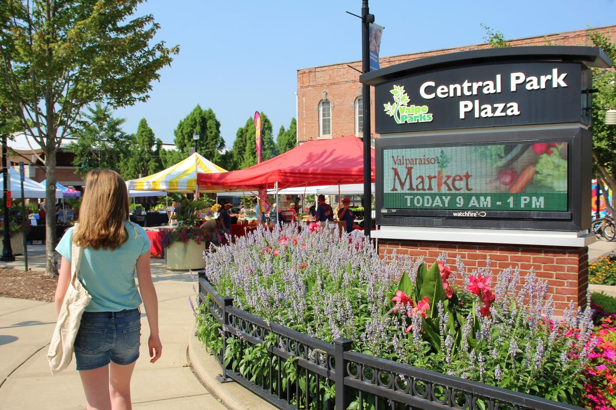 A young woman in a blue shirt and jean shirts walks toward tents in a market. A sign next to her is surrounded by flowers.
