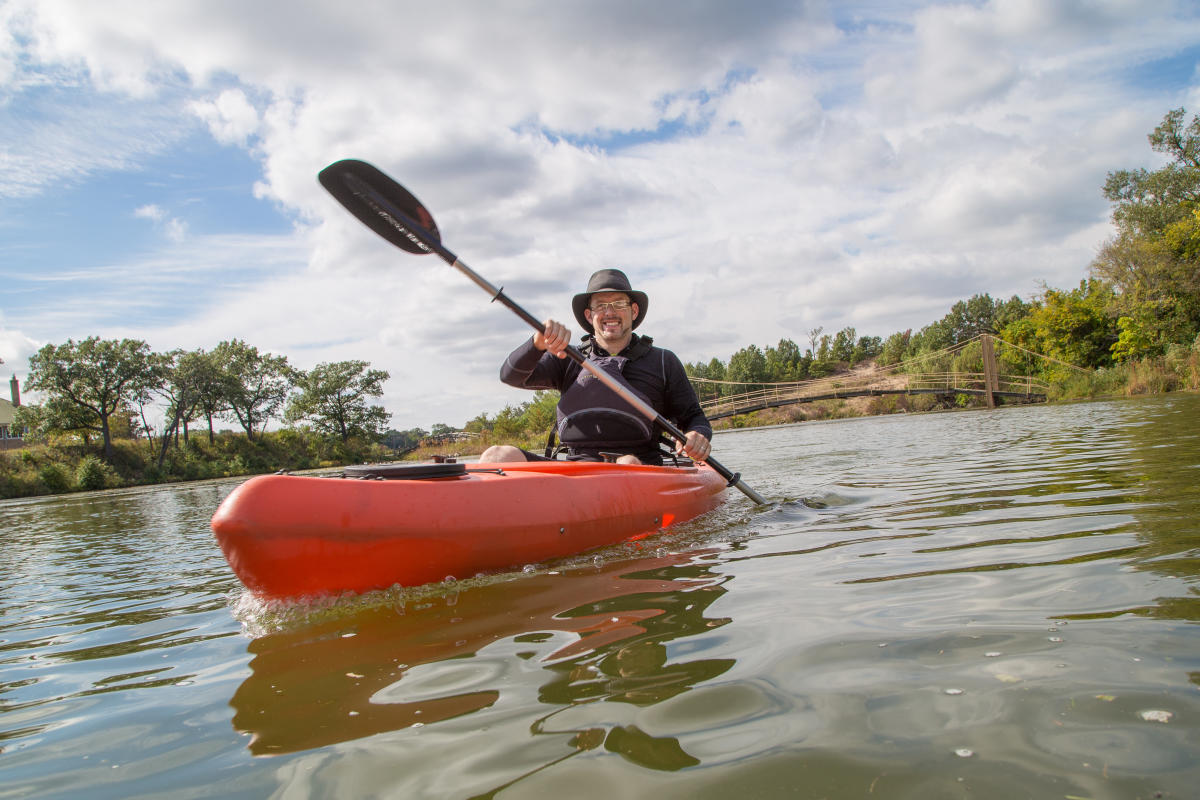 A man smiles as he sits in an orange kayak, paddling on the water. The sky is partly cloudy in the background.