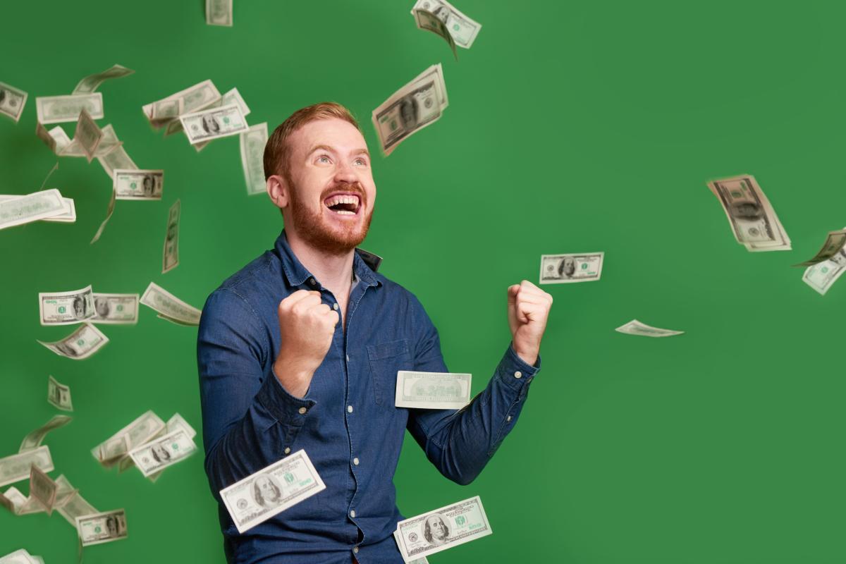 A man in a blue shirt looks up with a smile on his face as money falls from above him