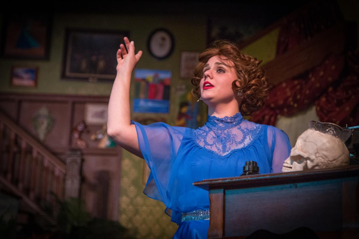 A woman in a bright blue dress acts on stage. Her had is raised in the air. A fake skull is on a pedestal in front of her.