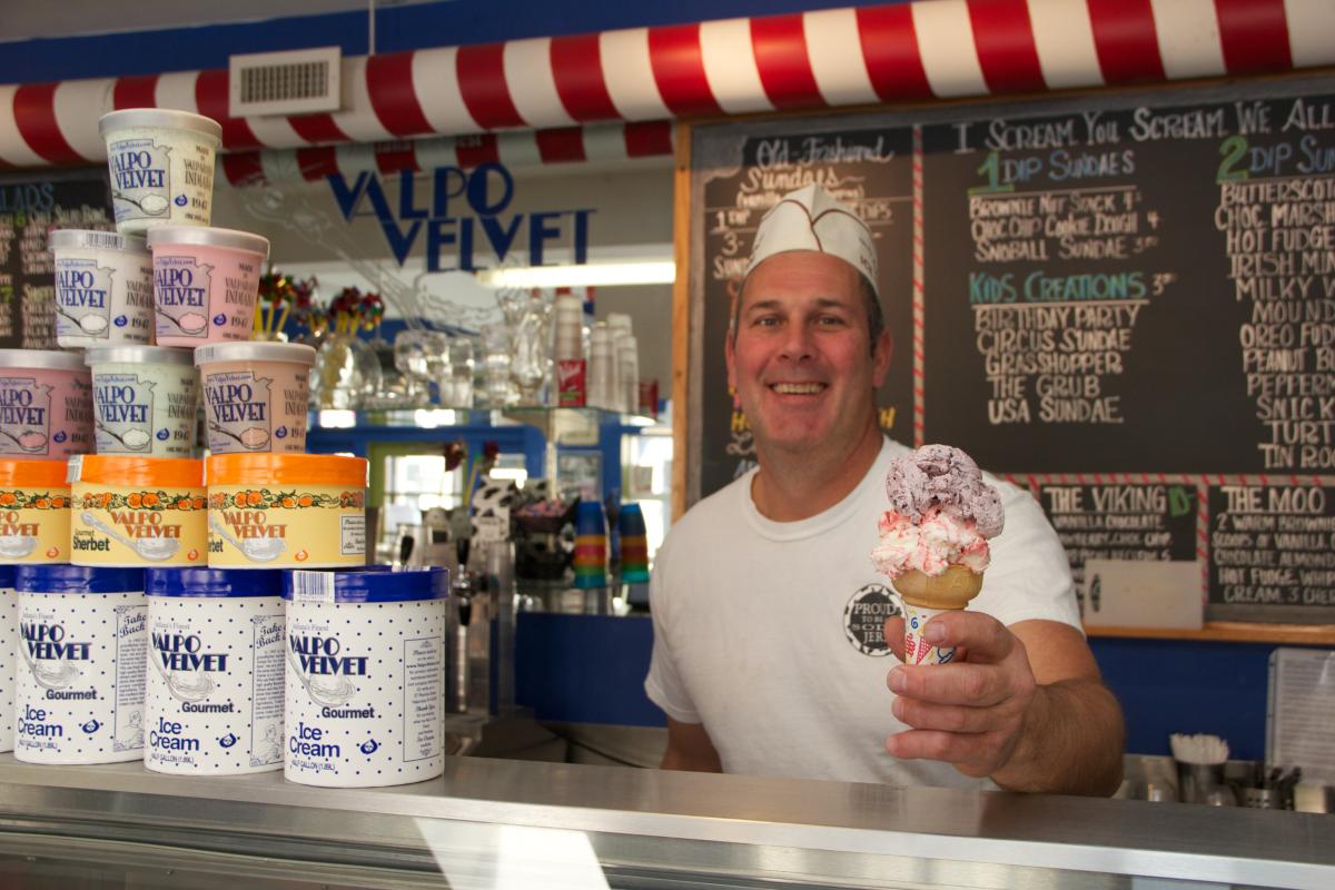 A smiling man hands a double scoop ice cream cone over the counter. Behind him is a chalkboard menu. To the left of him is a pyramid of ice cream pints.
