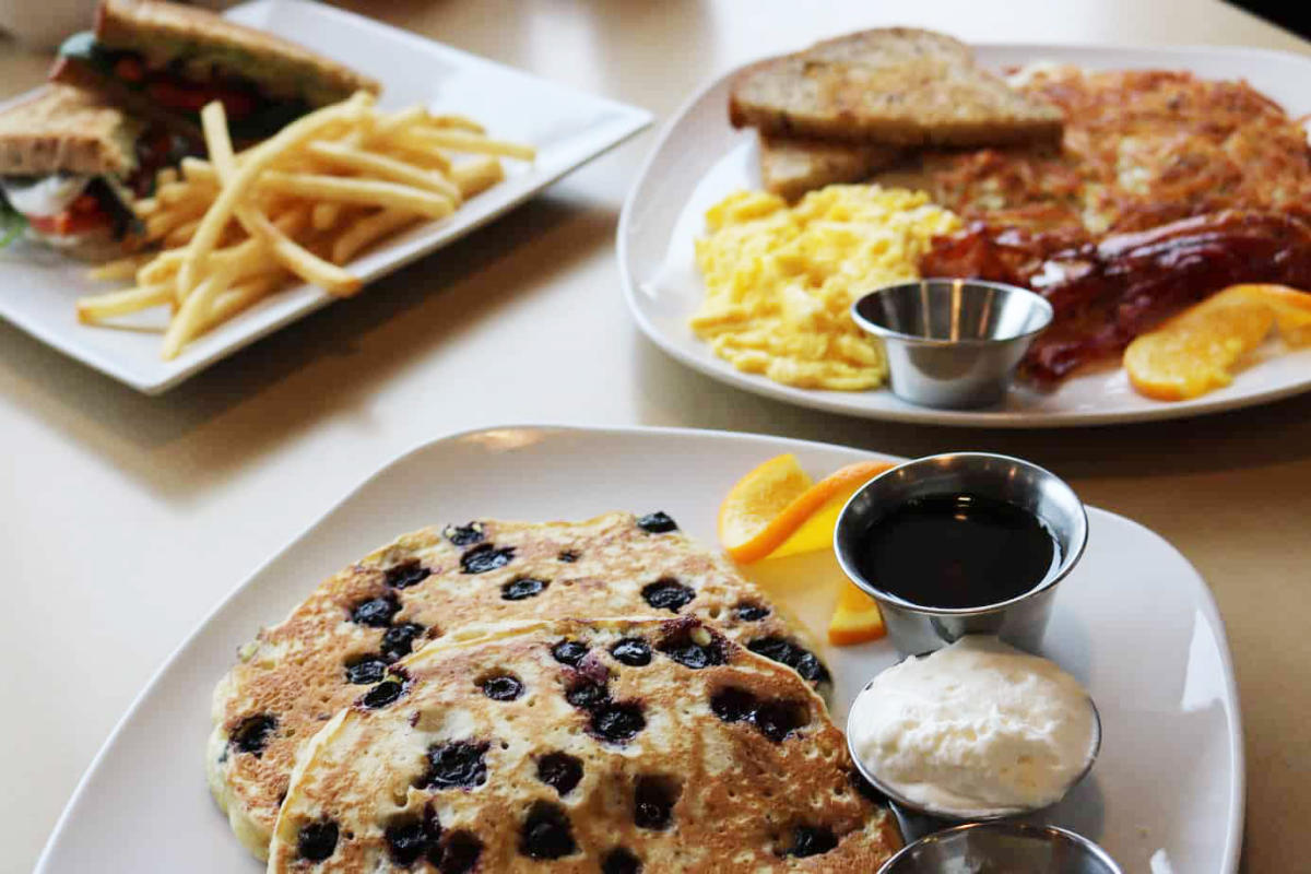 Three plates with cafe food sit on a table including blueberry pancakes, eggs and bacon, and a club sandwich with french fries.