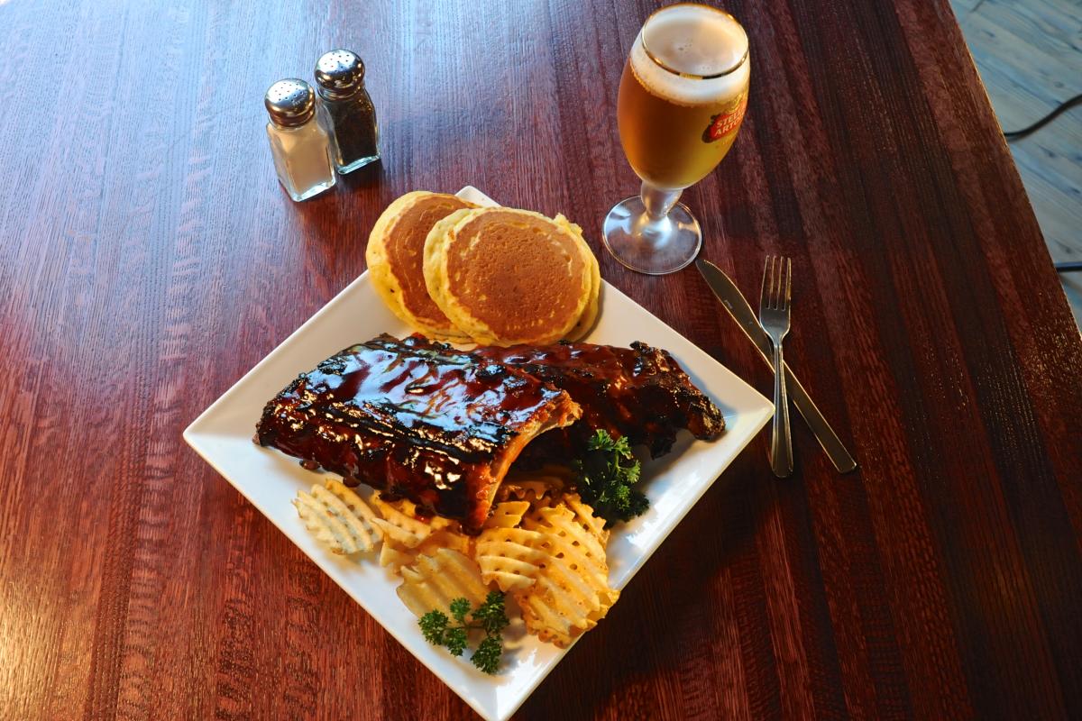 A square plate sits on a table with ribs, cross-cut potatoes, and pancakes. A beer, silverware, and salt and pepper shakers sit on the table.