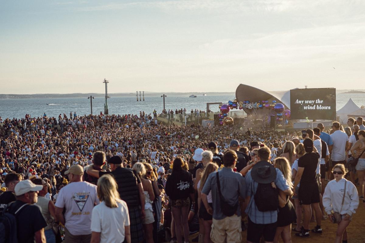 A large crowd gathers to watch a performance on the Bandstand Stage at Victorious Festival, with a calm sea in the background.