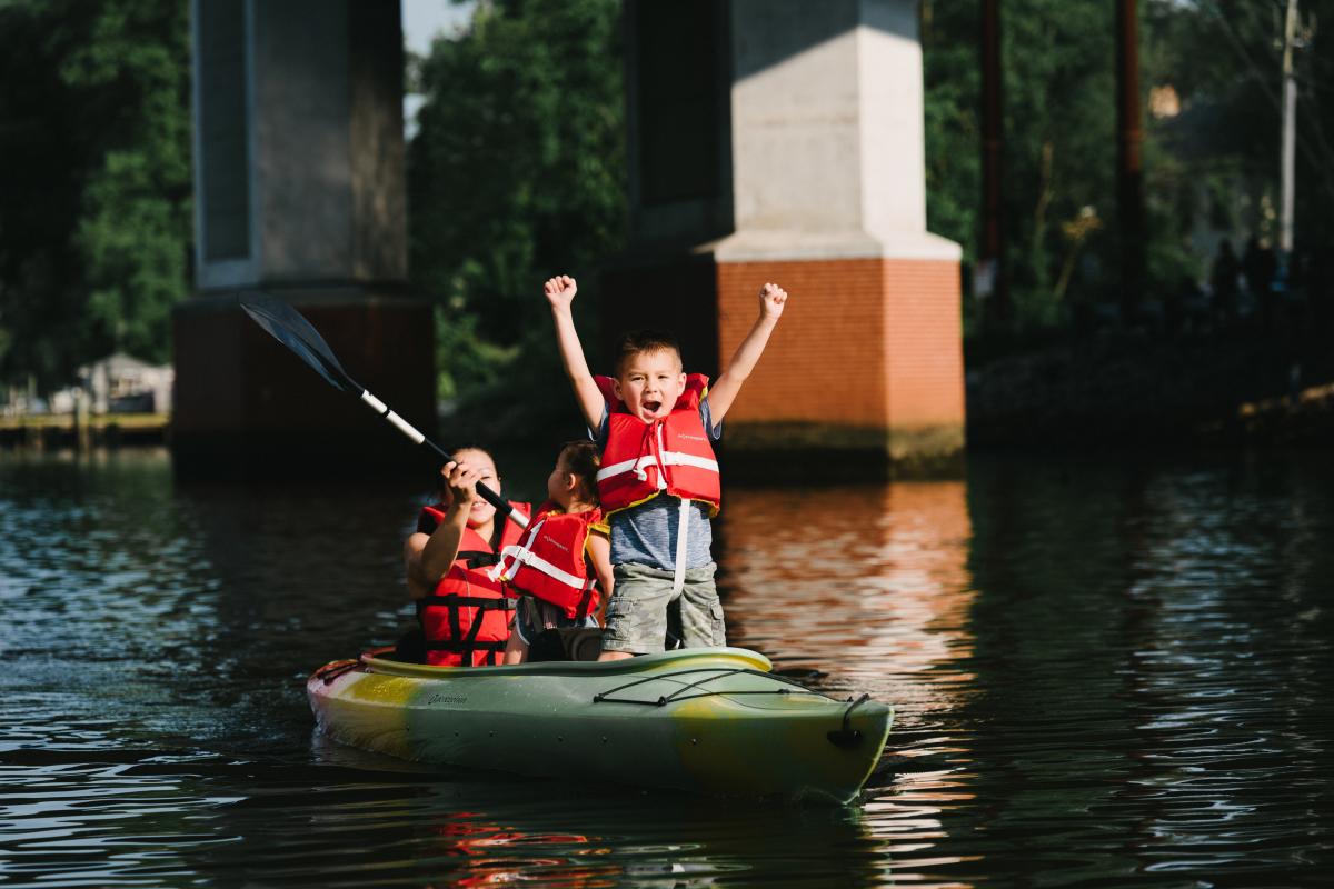 a woman and two children kayaking in a river, one of the children is holding both arms up excited