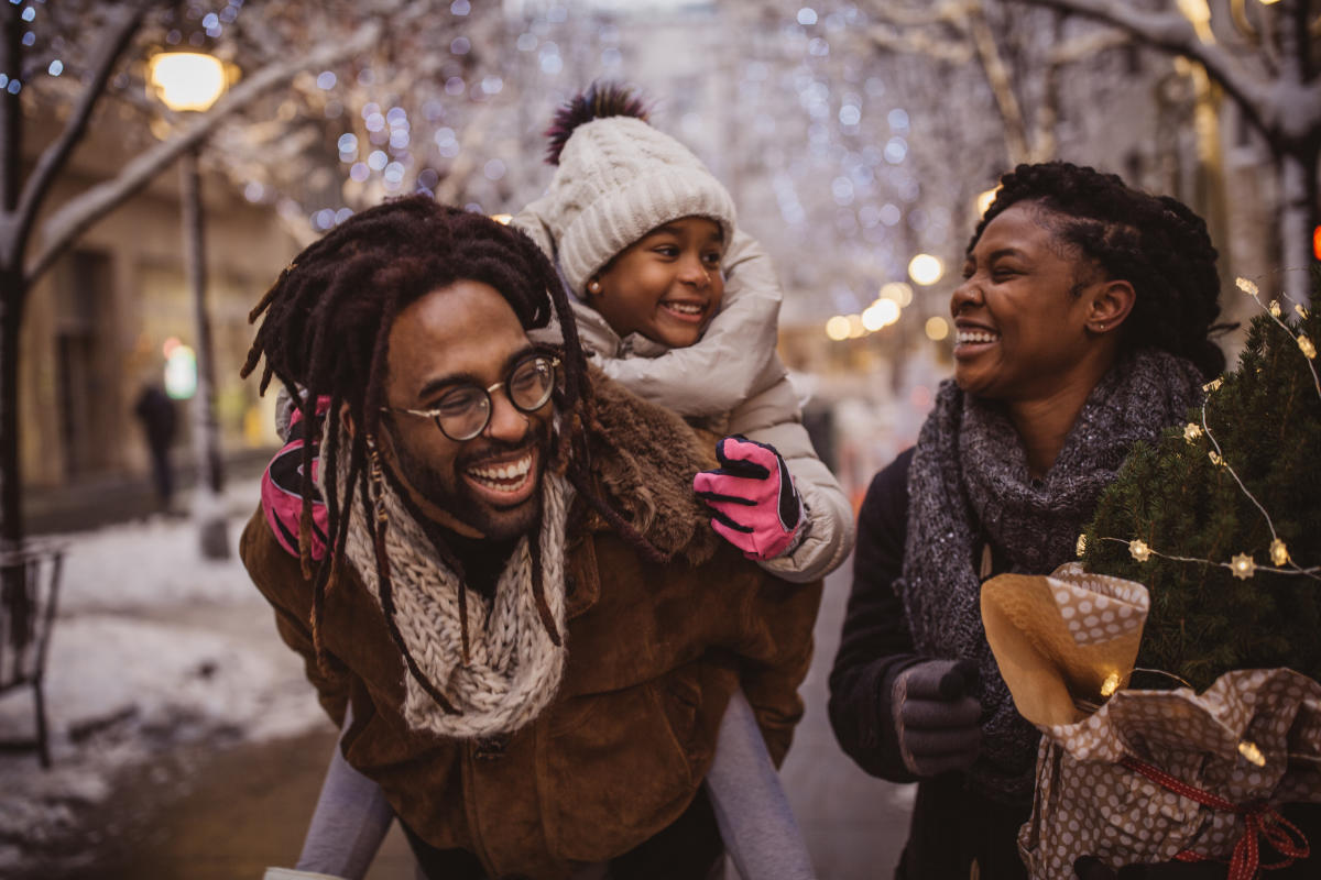 Black man carrying a child on his back with a woman beside them carrying a small christmas tree on snowy evening