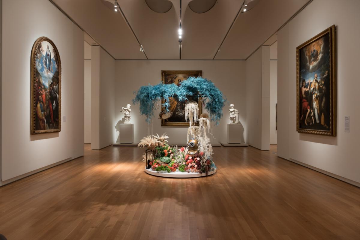 tall floral display sits in middle of museum gallery, empty aside from large-scale art on the walls