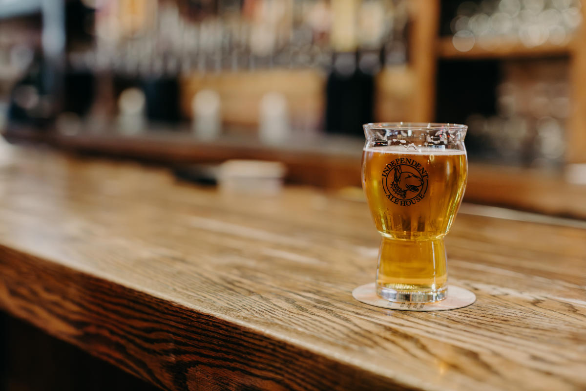 A glass of beer sits on a wooden bar countertop, with blurred shelves and bottles in the background. The setting feels warm and inviting.