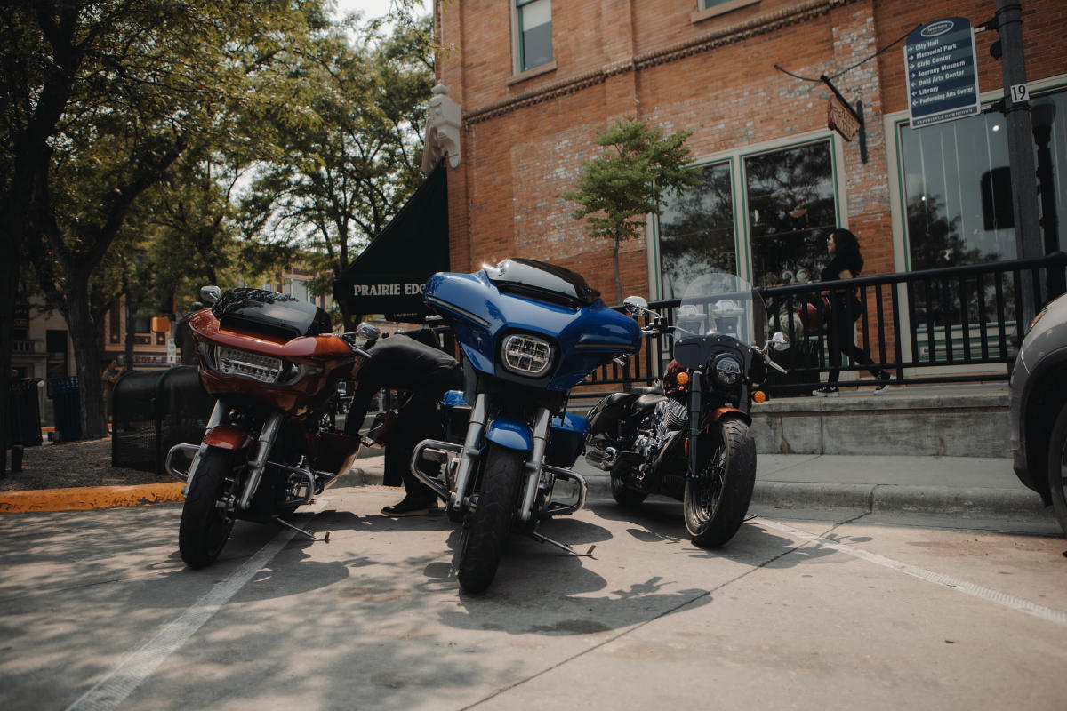 Three motorcycles, one red, one blue, and one black, are parked along the sidewalk, partially shaded by a tree, outside of Prairie Edge in Downtown Rapid City in mid afternoon.