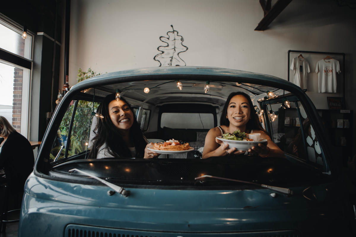 Two smiling women inside a vintage teal van, holding plates of food. String lights are visible, creating a festive atmosphere in the cozy setting.