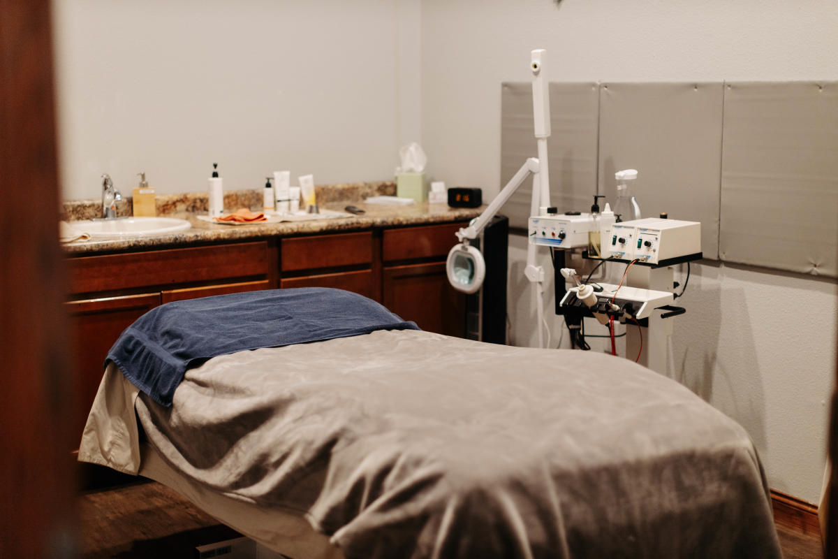 Calm spa room with a massage table covered in a beige blanket and blue towel. A countertop with skincare products and essential spa equipment nearby.