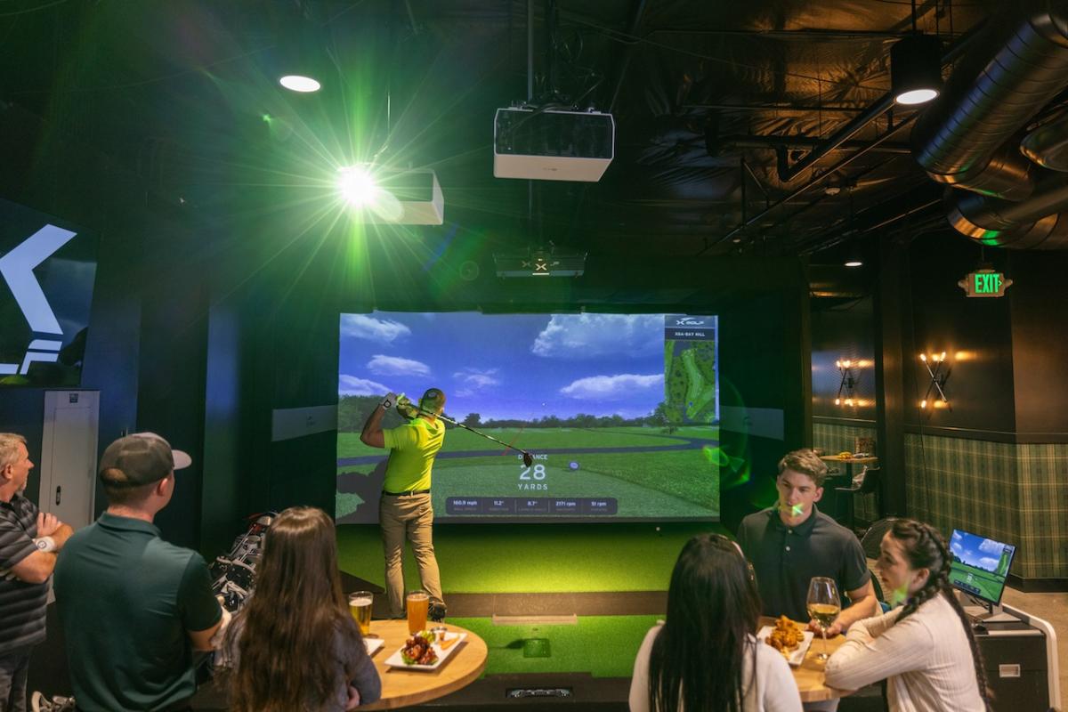 In the background, a man follows through on a golf swing on an indoor golf simulator while in the foreground, two groups of people are chatting, eating, and watching the golfer.