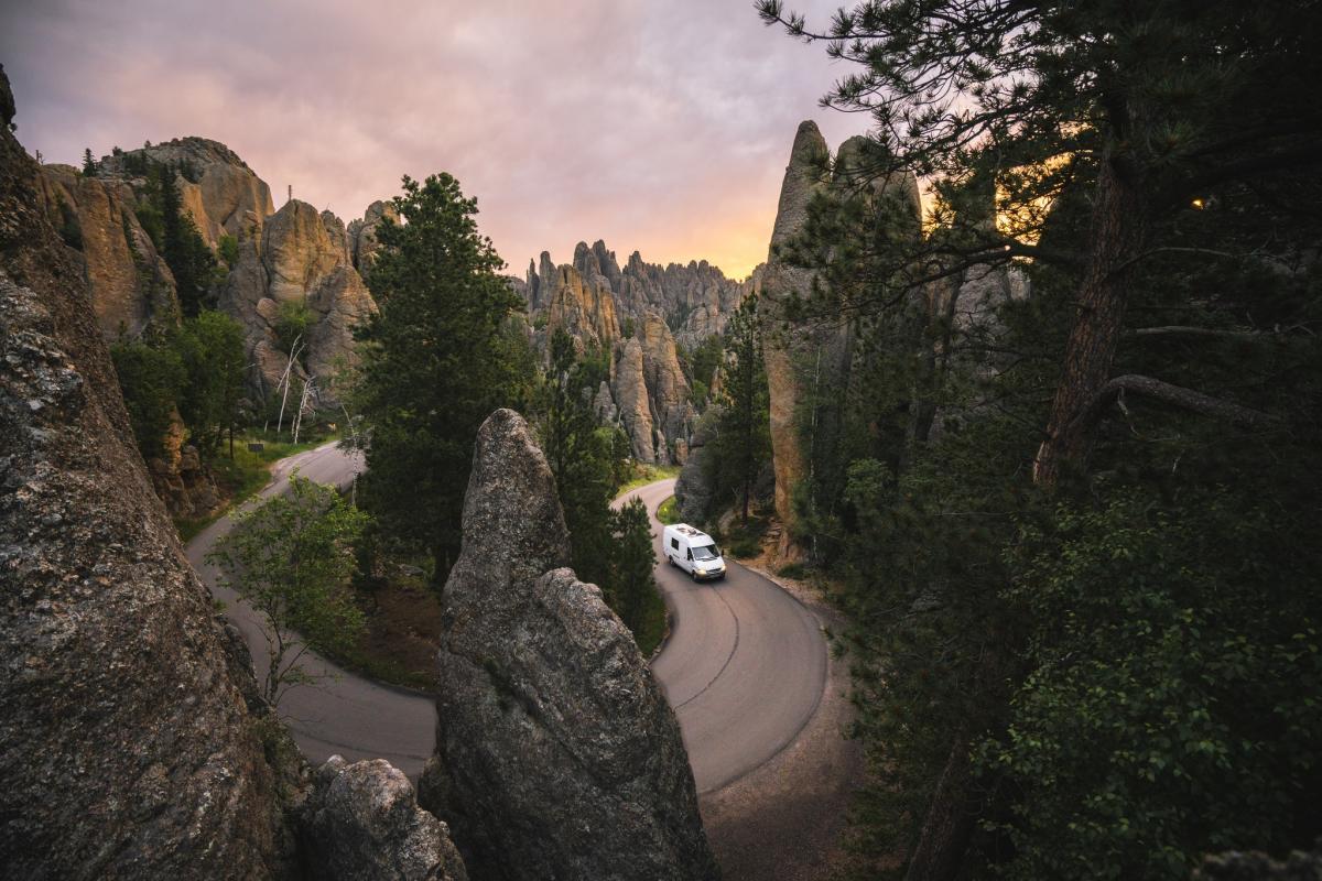 A white van navigates a winding road through rugged, rocky terrain surrounded by tall, jagged formations and lush trees at sunset, evoking adventure.