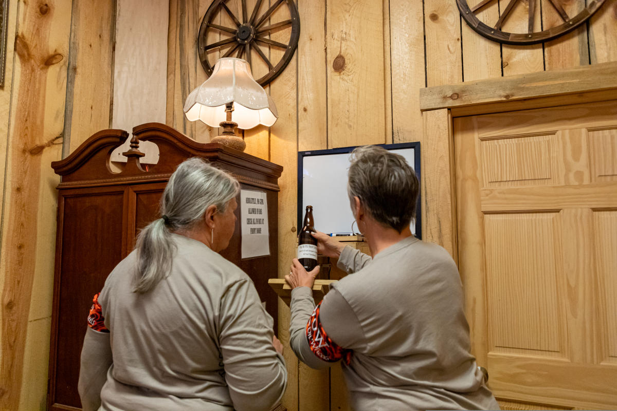 Two people in matching shirts stand in a rustic room with wooden walls. One holds a beer bottle, pointing to a TV. Wagon wheels and an antique cabinet create a cozy, vintage vibe.