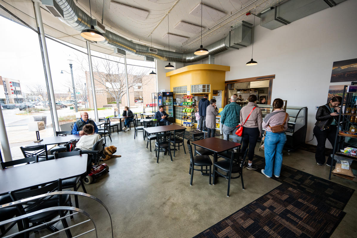 Modern cafe scene with several customers. People sit at tables by large windows and others queue at a counter.