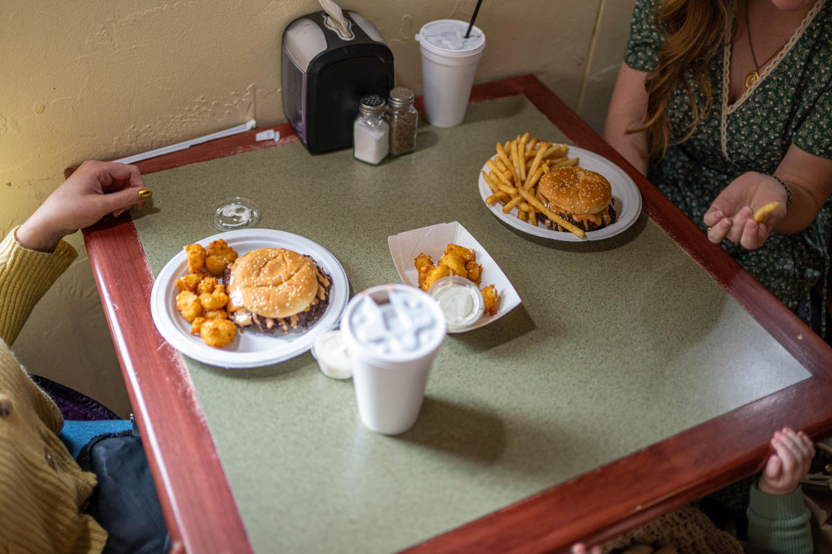 Two women sit at a table with cheeseburger meals sitting in front of them. One holds a French fry in her hand. Two Styrofoam drink cups sit beside their plates.