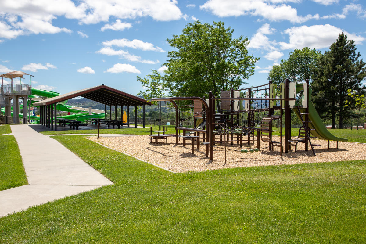Playground and picnic shelter at Horace Mann Park in Rapid City on a sunny day. A modern climbing structure and slide sit beside paved walking paths and open green space, while the waterslides of the Horace Mann Pool provide a colorful backdrop against the Black Hills foothills.