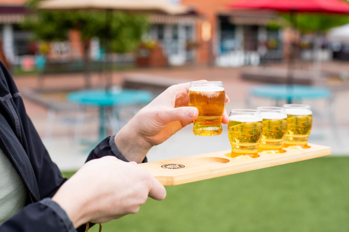 A person holding a flight of four small glasses of beer while picking up the first glass.