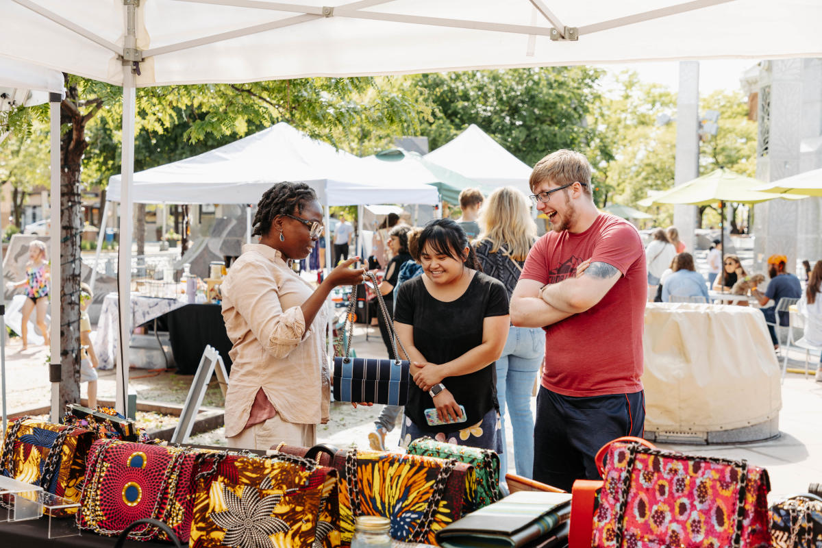 A male and a female shoppers laugh with a saleslady while browsing colorful bags under a tent at Main Street Square.