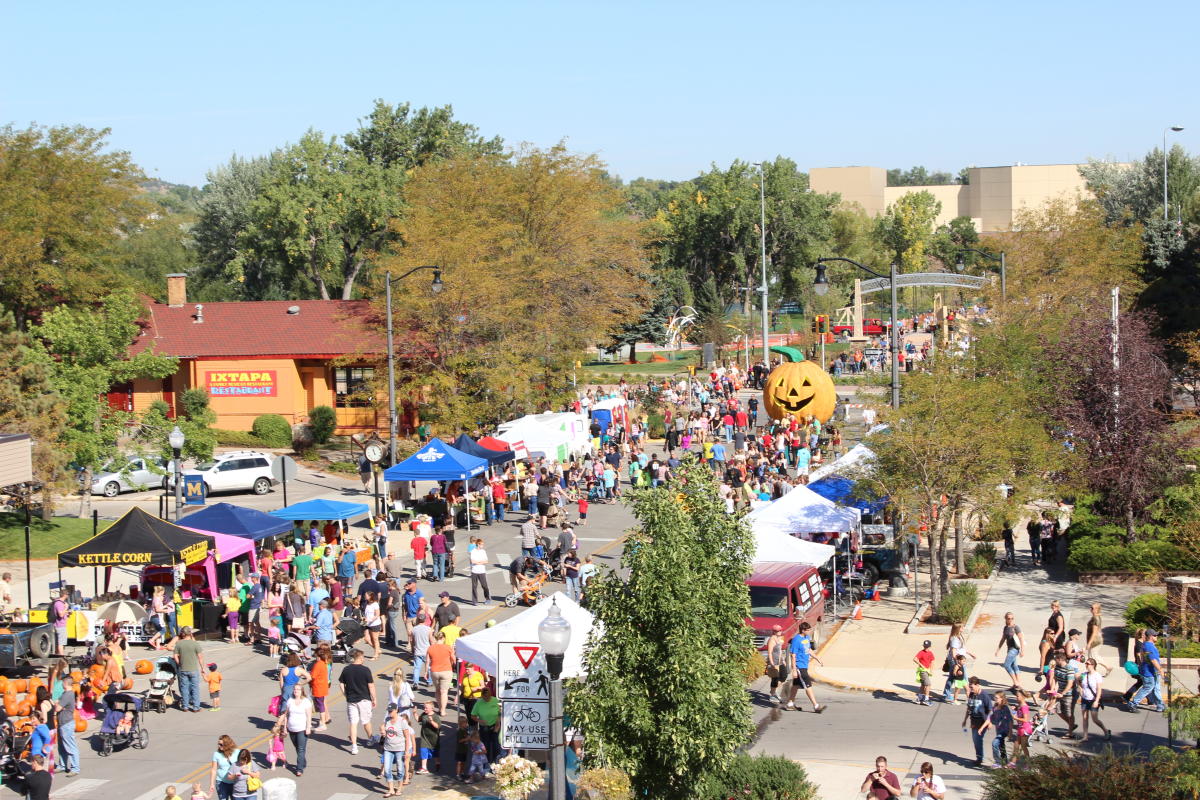 A bustling fall festival with colorful vendor tents, crowds of people, and a large inflatable pumpkin. Trees with autumn foliage and a sunny sky convey a festive, lively atmosphere.