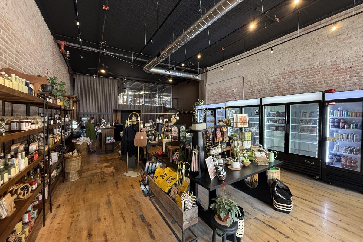 Spacious store interior with wooden floors and brick walls. Shelves are stocked with various goods, including plants and signs. Refrigerators line the right wall. Warm, inviting ambiance.