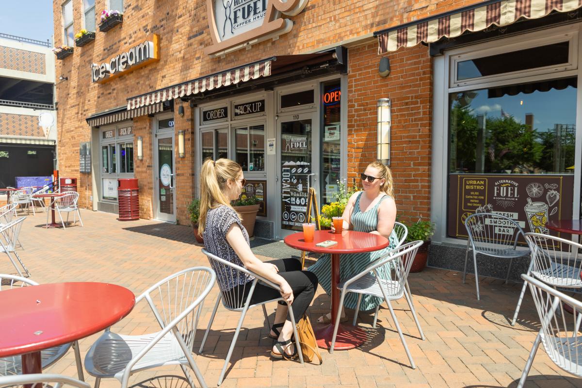 Two women sit at a small bright red table on the patio of Alternative Fuel Coffee House in the bright sunshine.