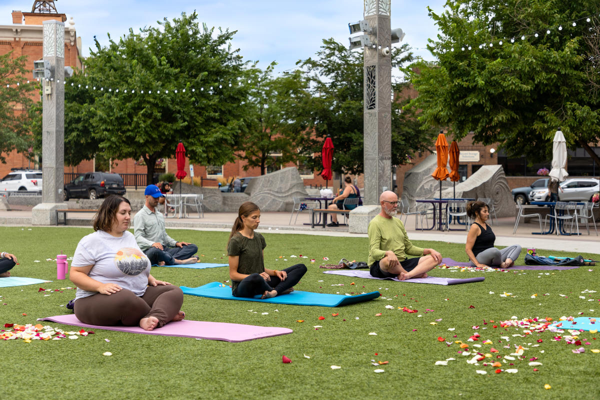 A group of people meditate on yoga mats, surrounded by scattered rose petals. The scene is serene, with trees and a brick building in the background.