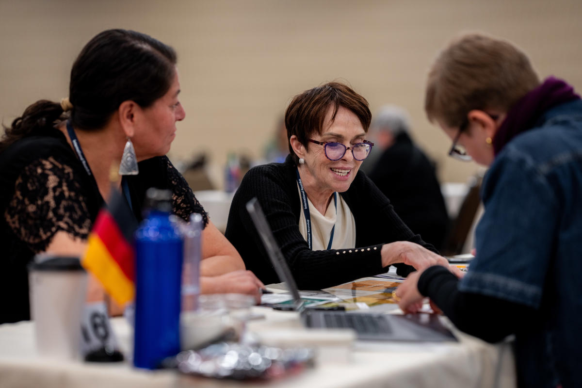Three people engaging in a lively discussion at a table with laptops and papers. A small German flag and a blue water bottle are visible. The mood is collaborative.