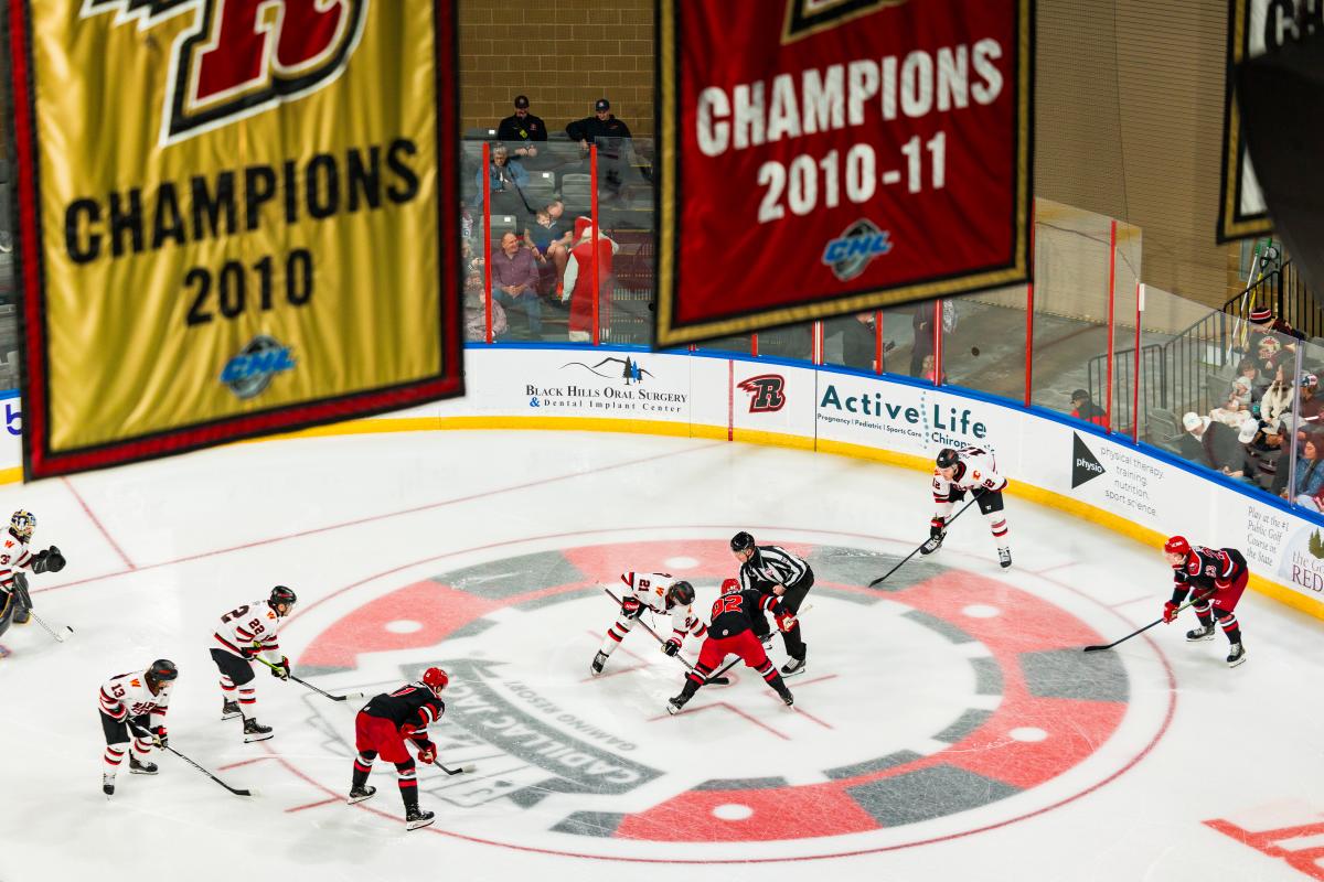 Hockey players from two teams line up for a faceoff while a referee drops the puck, with fans in the stands behind the glass and large championship banners hanging overhead in the arena.