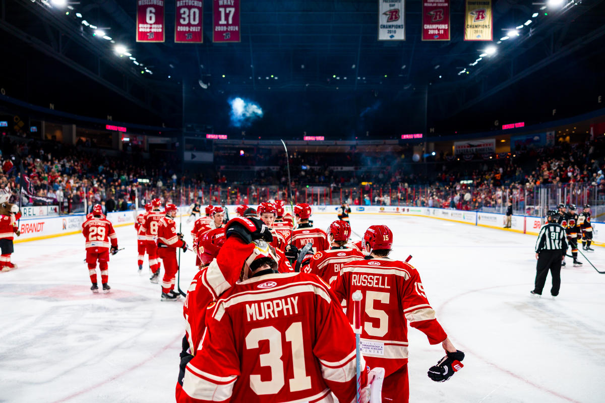Hockey players in red jerseys skate off the ice, celebrating a victory. A cheering crowd fills the arena, banners overhead. Energetic, triumphant scene.