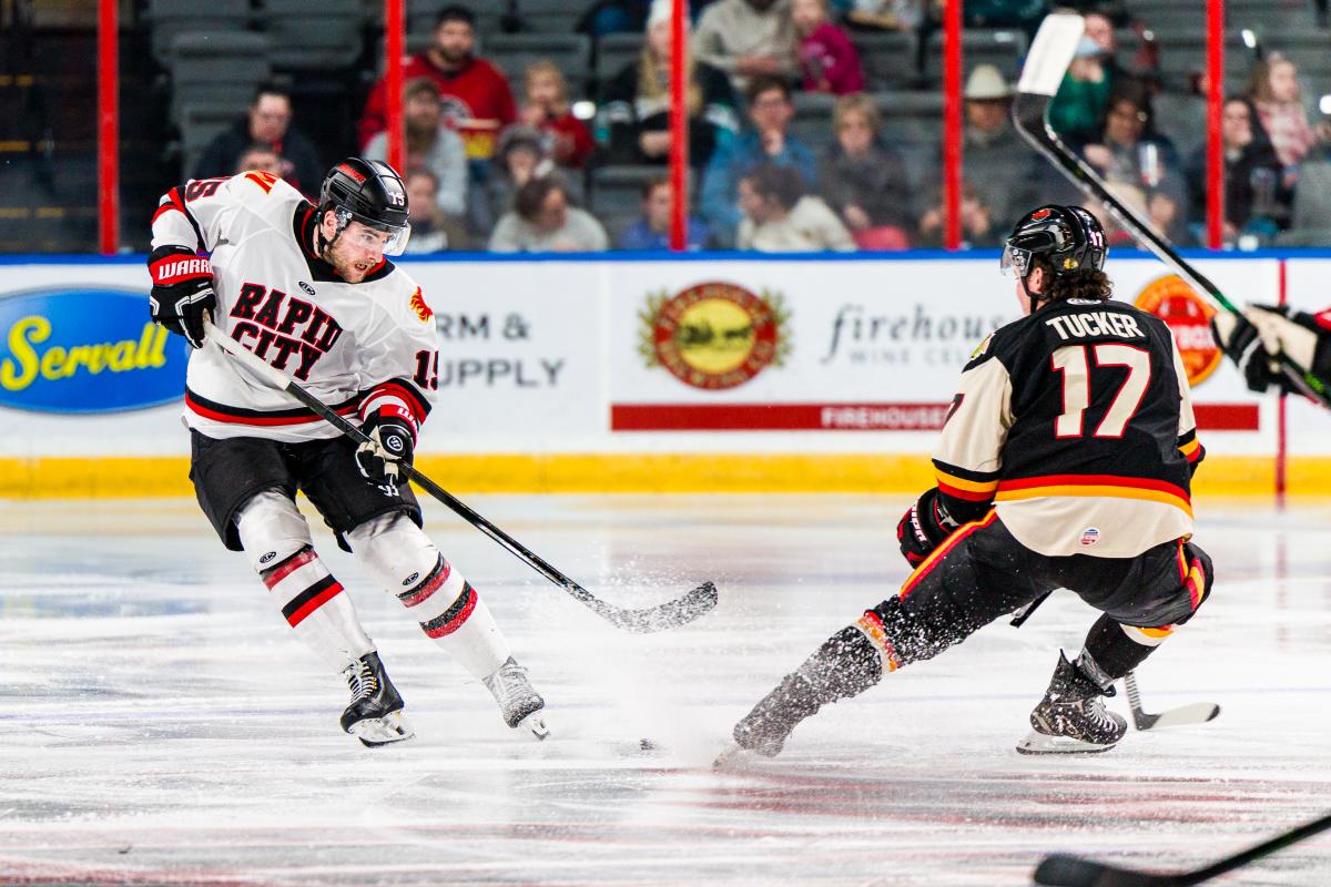 Two ice hockey players face off mid-play on the rink—one in a white Rapid City jersey skating forward with the puck, and one in a black jersey defending—while ice sprays from their skates and spectators watch from the stands behind the boards.