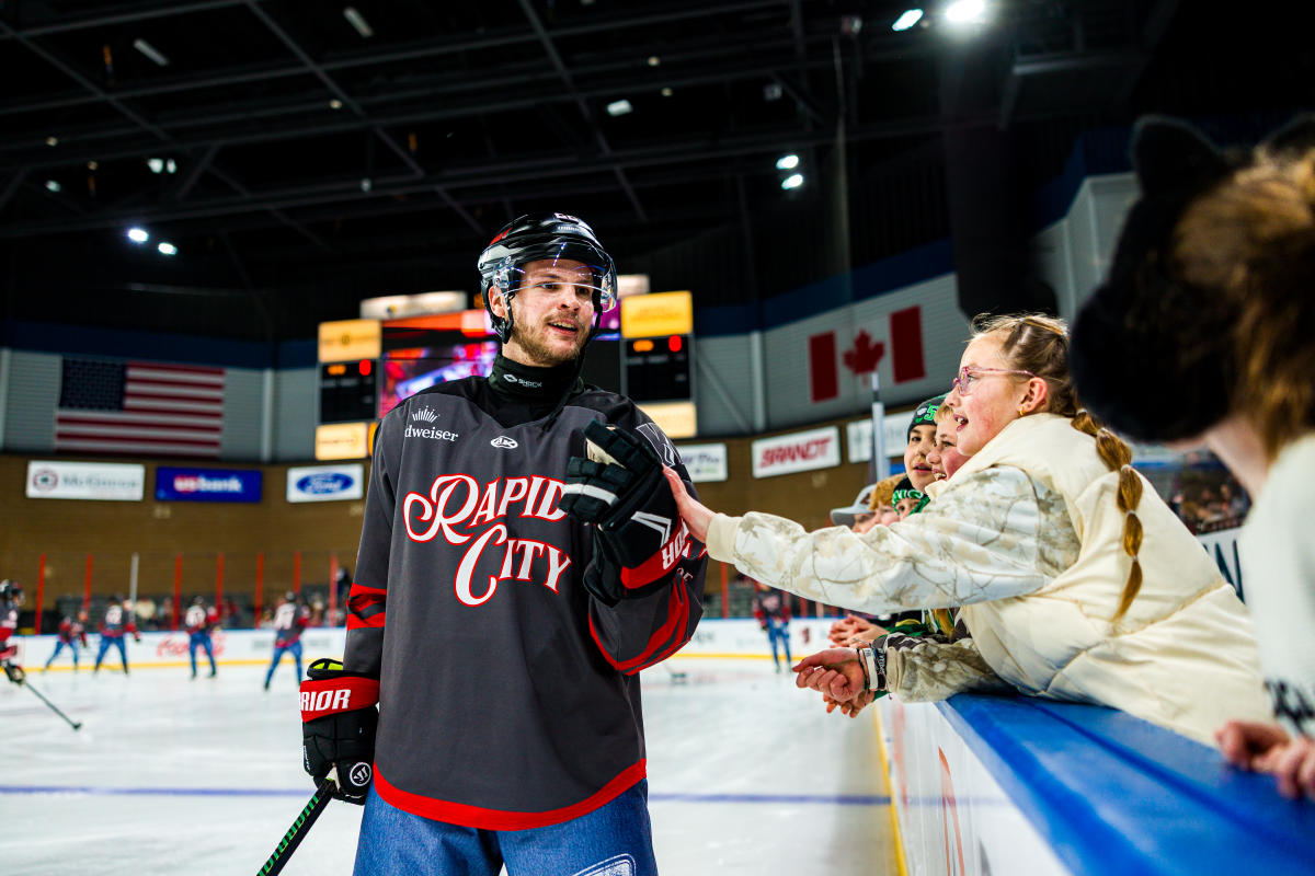 A hockey player in a charcoal gray Rapid City jersey fist bumps young fans who are watching warm ups on the player bench.