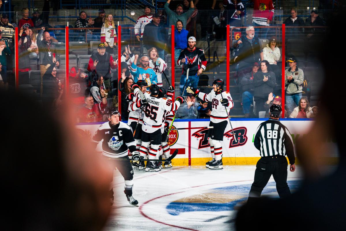 Hockey players in white jerseys gather for a group hug after scoring a goal. Fans behind the boards stand and celebrate with the team.