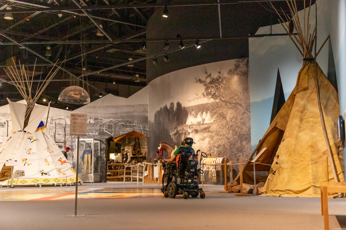 A man in a wheelchair makes his way through the Lakota history section of the Journey Museum, with two replica teepees on either side of a large walk-through exhibit.