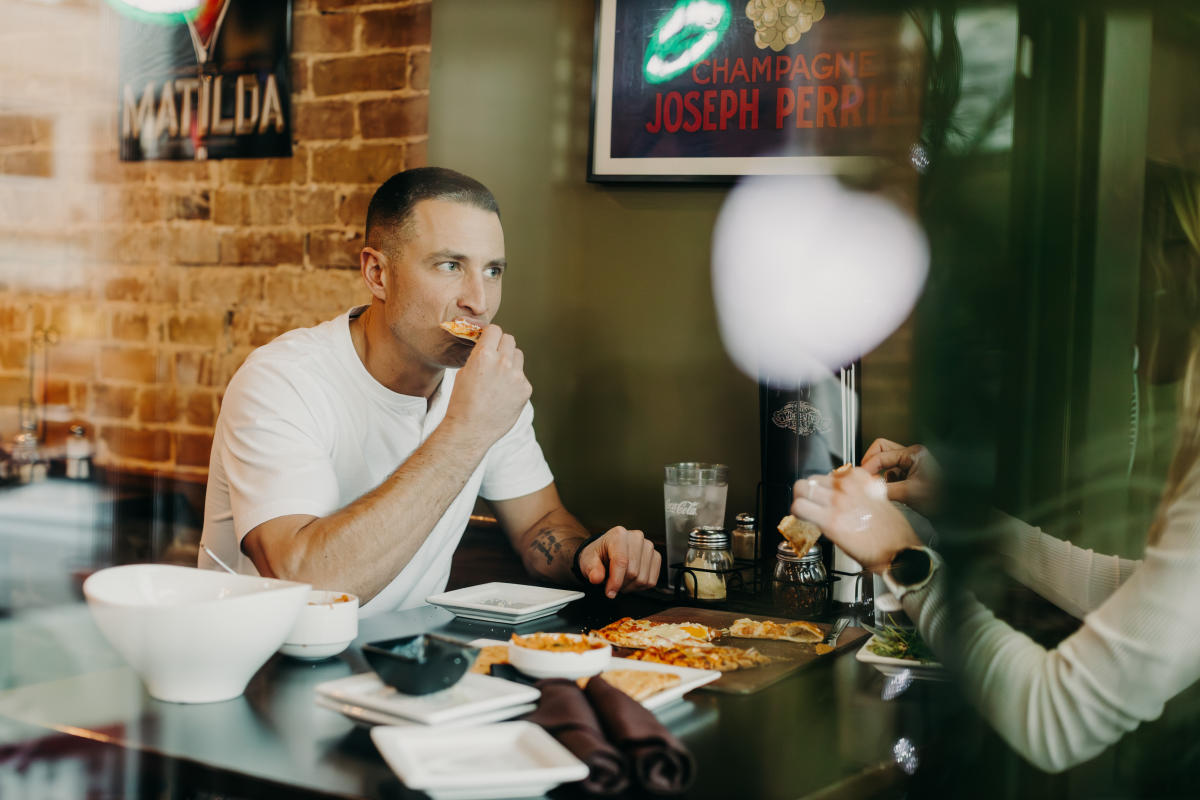 A man enjoys pizza at Independent Ale House, seated at a table with a spread of food. Exposed brick wall and vibrant posters add charm.