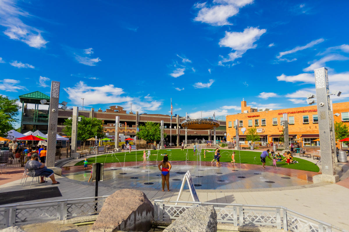 Children play in the interactive fountains at Main Street Square in downtown Rapid City under a bright blue sky with scattered clouds. Families relax and gather around the splash area, with shops, restaurants, and the Main Street Square pavilion surrounding the open plaza.