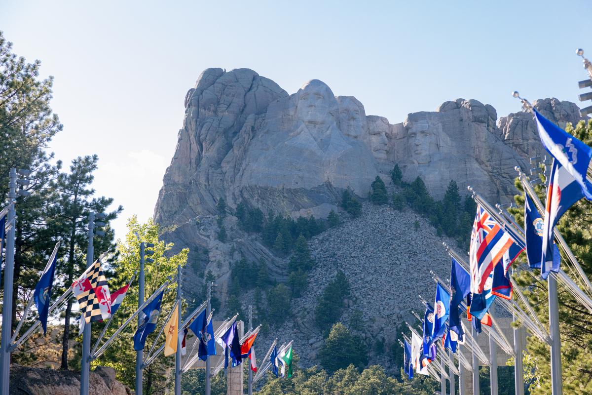 Mount Rushmore in bright daylight with clear blue sky. Flags from various states line the walkway, adding colorful contrast to the rocky landscape.