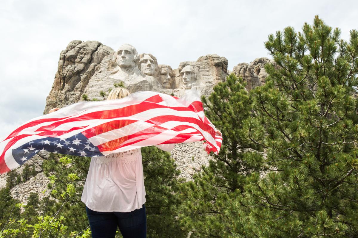 A woman holding an American flag behind her faces away from the camera toward Mount Rushmore, visible in the upper half of the photo under a mostly cloudy sky.