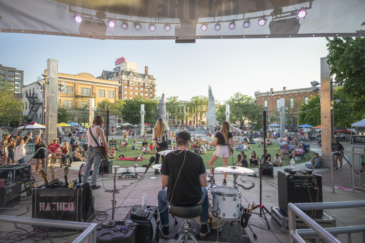 Outdoor concert scene with a band performing on stage, viewed from behind the drummer. Audience relaxes on grass, under a clear sky, enjoying the music.