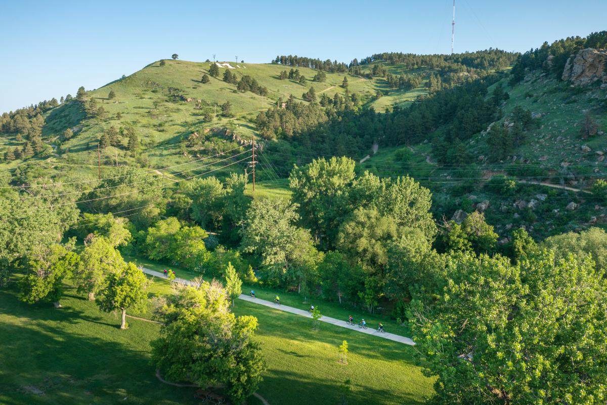 Aerial view of cyclists riding along a pathway at Hanson-Larsen Memorial Park amidst lush green trees and grassy hills. The scene is serene, with clear blue skies and a feeling of tranquility.