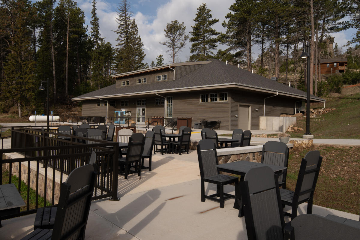 Outdoor patio seating at the Sylvan Lake General Store in Custer State Park, featuring black tables and chairs on a spacious concrete terrace. The lodge-style building sits among towering pine trees and granite outcroppings, creating a scenic Black Hills setting near Sylvan Lake.