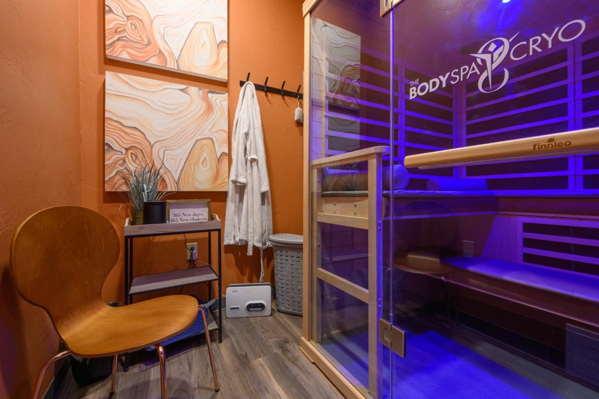 A cozy spa room with orange walls, abstract art, a wooden chair, a hanging robe, and a glowing blue-lit cryotherapy chamber labeled "Body Spa Cryo."