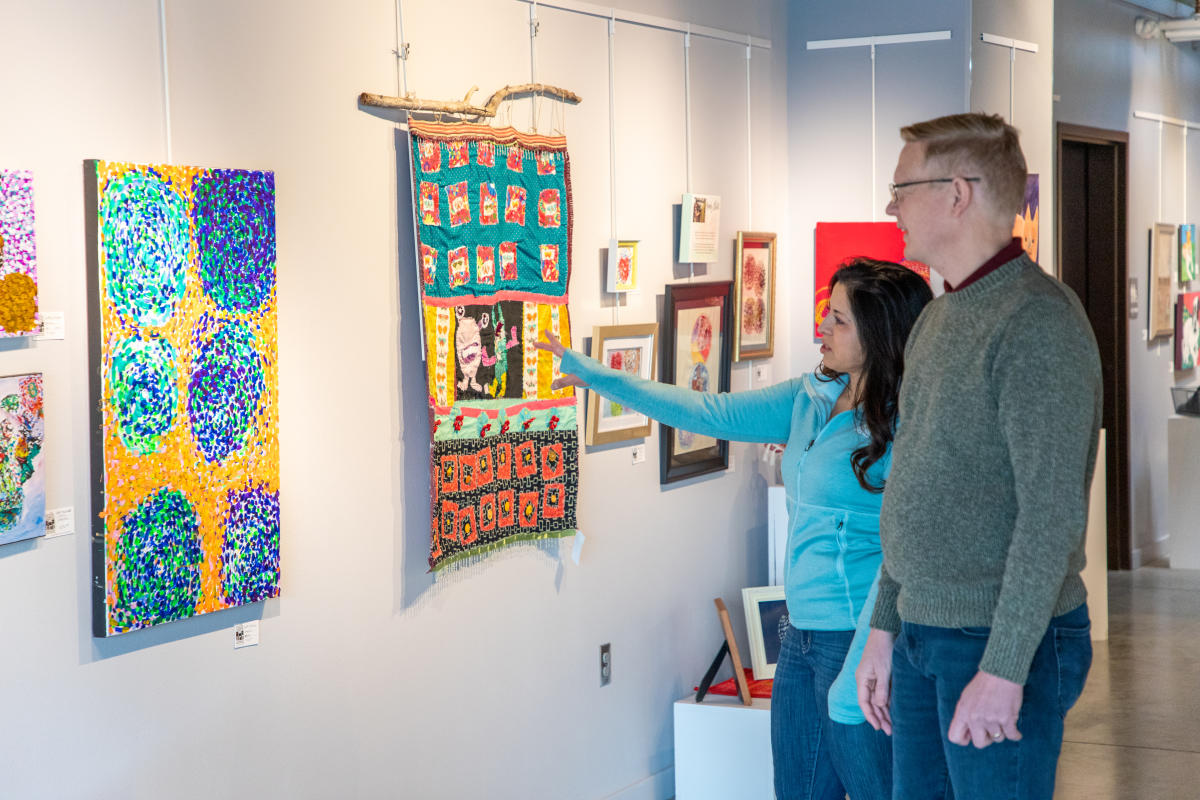 Two people observe colorful artwork in a gallery, with vibrant paintings and textiles on the wall. The atmosphere is engaging and thoughtful.