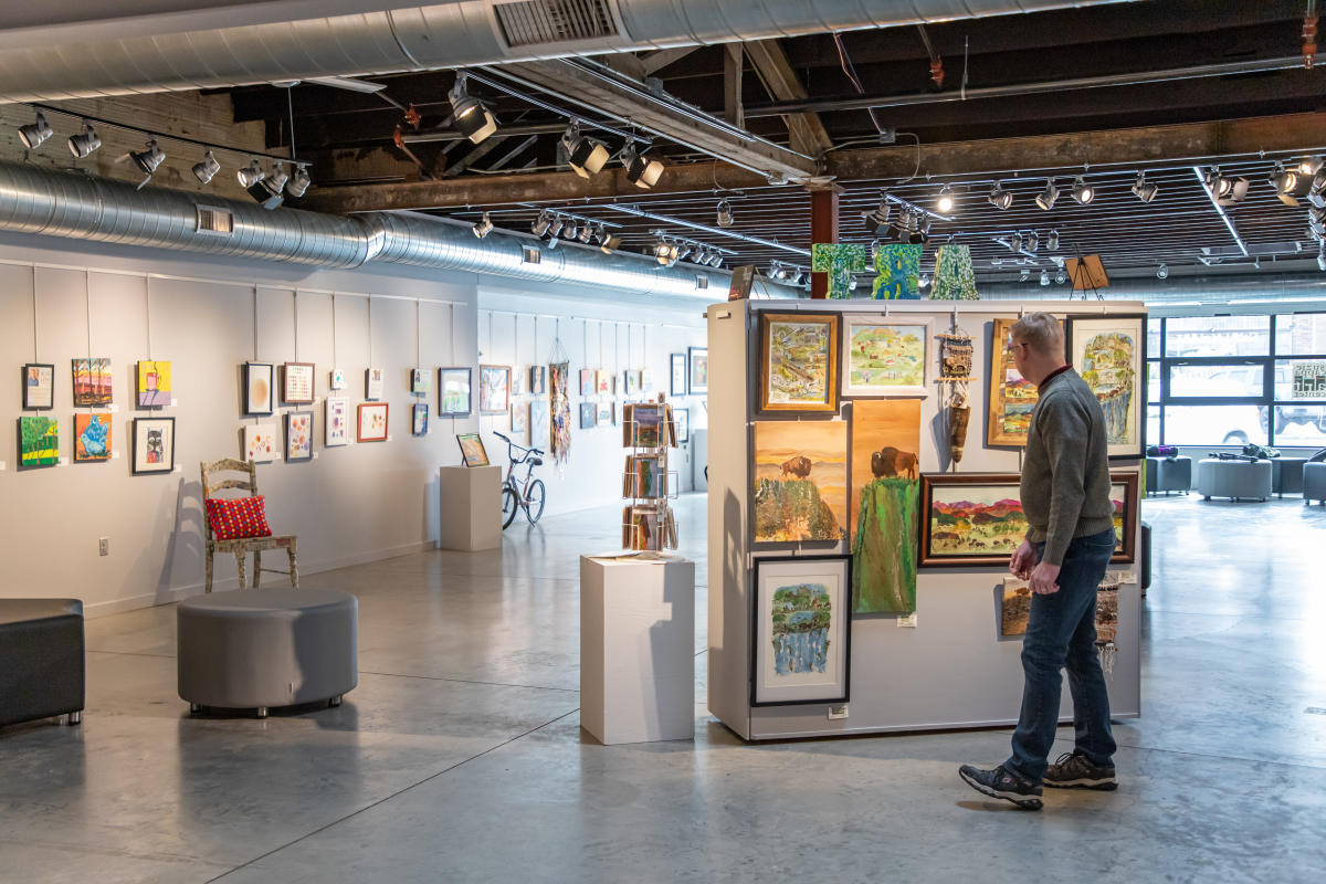 An art gallery with paintings displayed on walls and panels. A person in casual attire observes a panel of colorful landscape artworks, creating a serene and contemplative atmosphere.