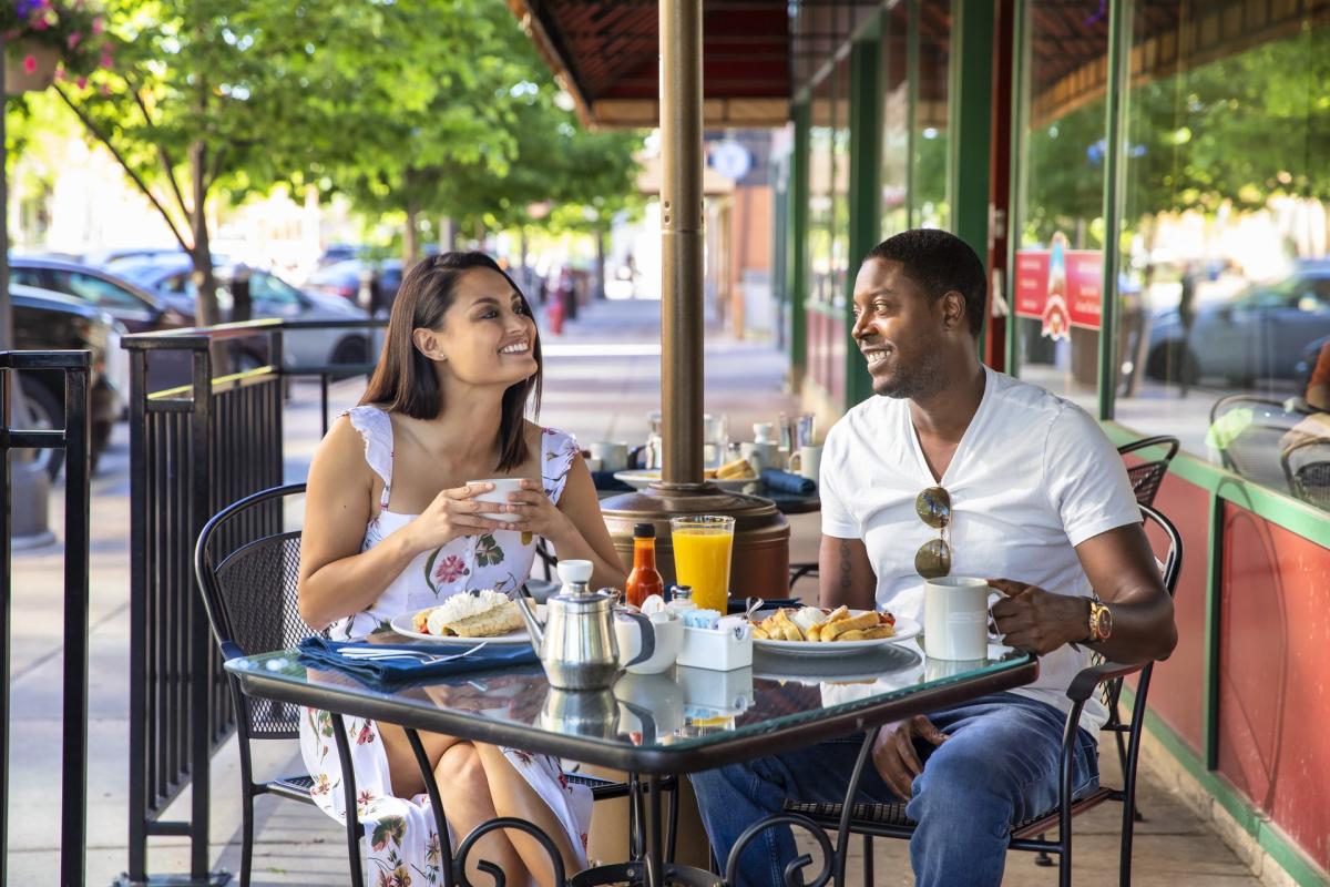 A smiling couple enjoys an outdoor brunch at Tally’s Silver Spoon. The woman in a floral dress holds a cup, while the man in a white shirt smiles back, creating a warm and cheerful atmosphere.