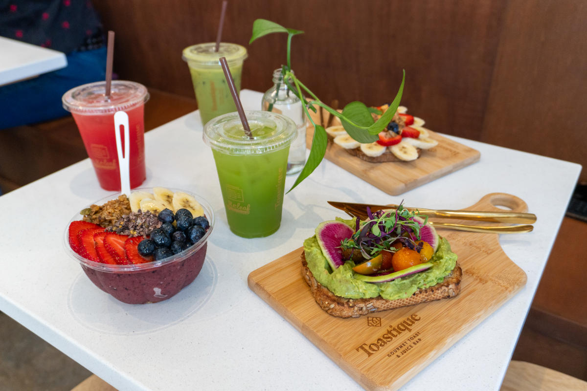 Spread of avocado toast, acai bowl, green juices, and smoothies on a white table at Toastique.