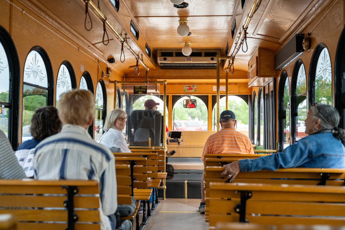 Passengers ride inside the City View Trolley, seated on wooden benches along the polished wood interior, with large arched windows, brass handrails, and vintage-style lighting. A trolley operator and several passengers are visible toward the front.