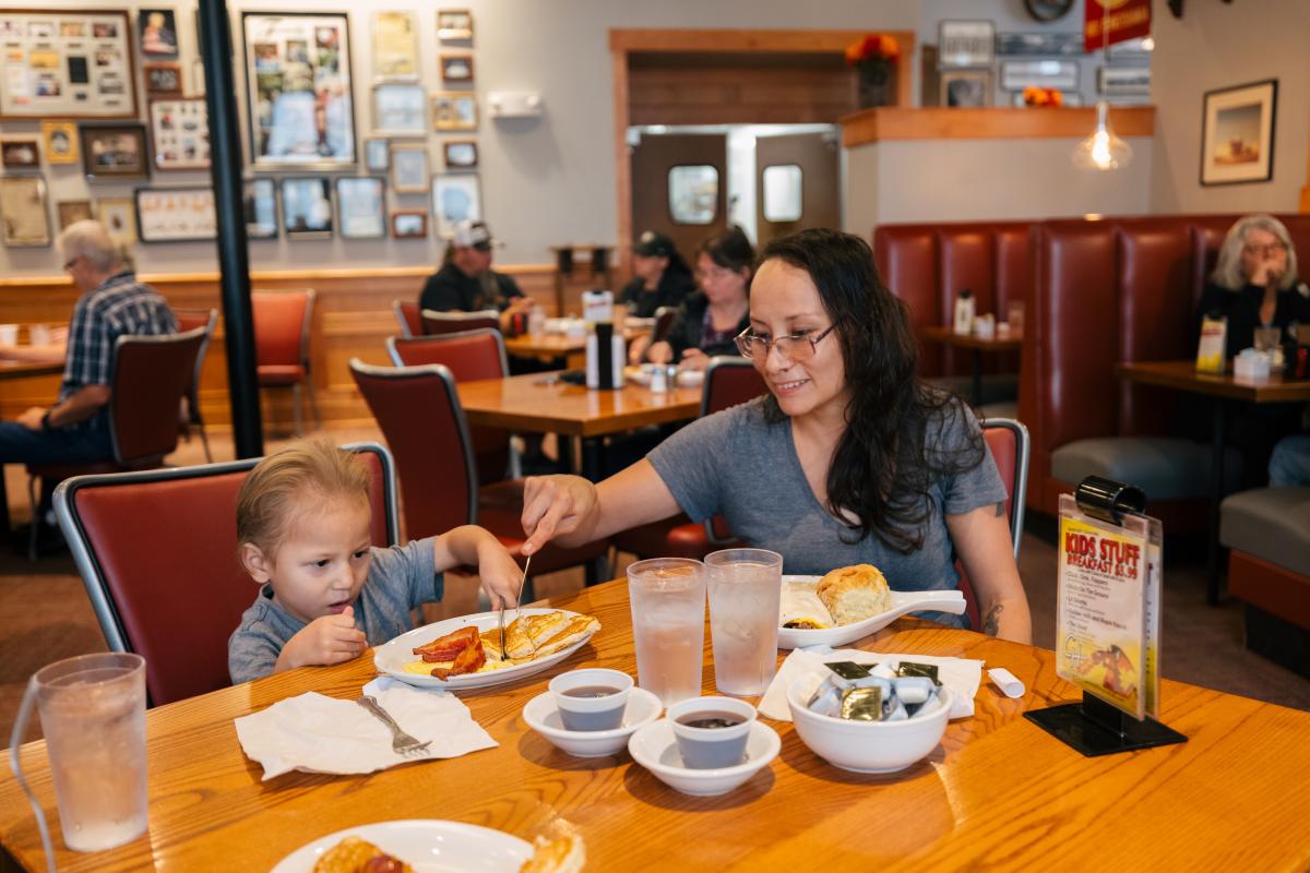 A mother and young child enjoy breakfast in a cozy diner. She helps him with a fork, surrounded by warm decor and other diners. The mood is cheerful.