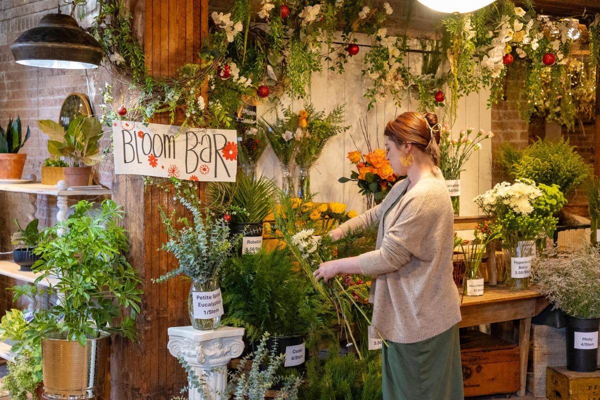 A woman arranges flowers at a "Bloom Bar" inside of Victoria’s Garden decorated with plants, bouquets and greenery. Various floral arrangements are displayed on tables and shelves.