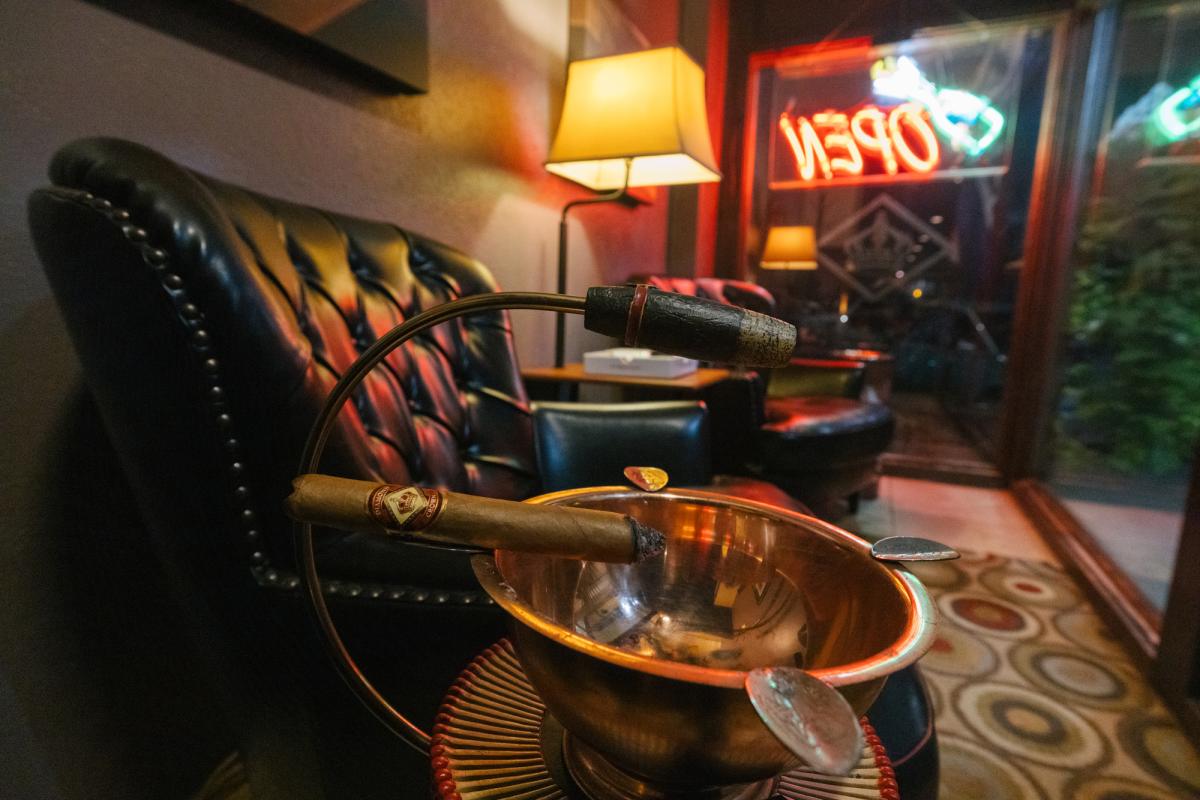 Premium cigar resting on a stand above a copper ashtray inside Tinder Box Diamond Crown Cigar Lounge in downtown Rapid City, with leather seating and a glowing neon open sign in the background.